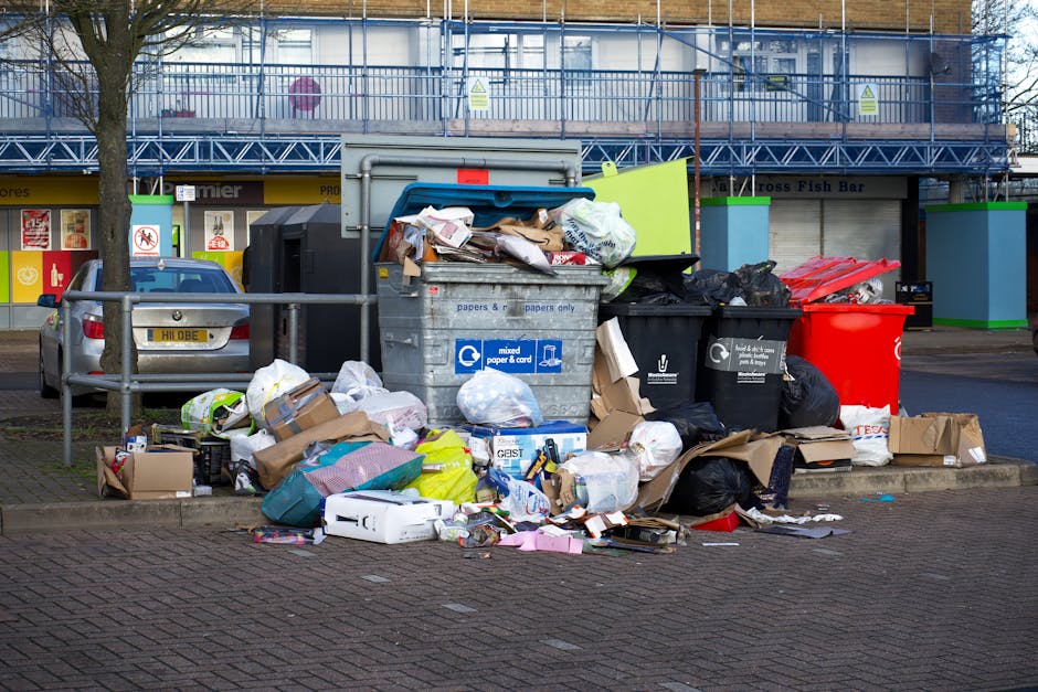 A pile of discarded cardboard boxes, some flattened and others partially intact, along with plastic packaging and small paper scraps, are gathered at the base of an external brick wall and next to a t
